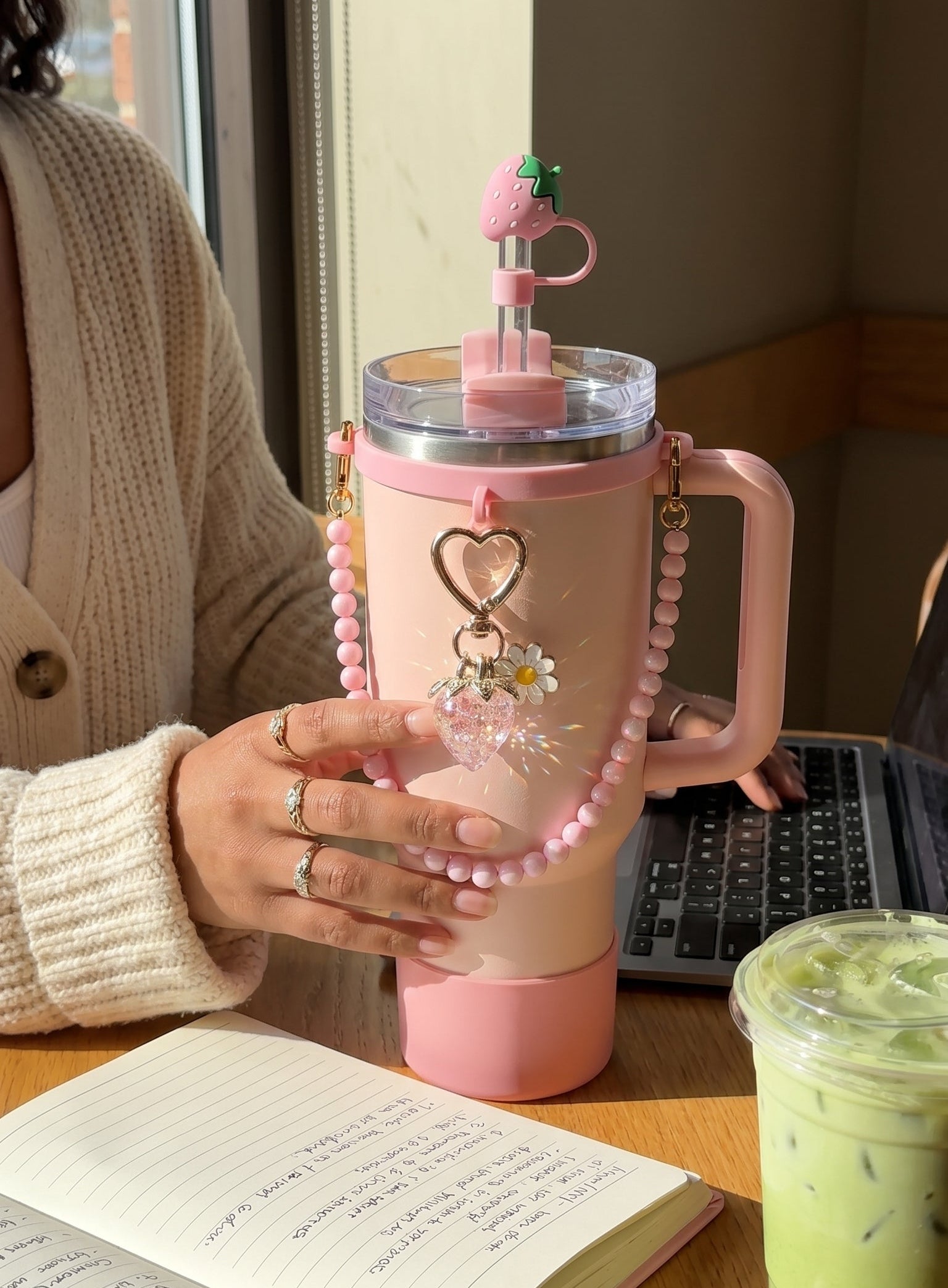 Person holding a pink tumbler with decorative elements on a desk next to an open book and a green drink.